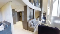 This contemporary lobby showcases refined architectural design with an emphasis on clean lines and upscale finishes. The space commands attention with its soaring double-height ceilings, enhanced by a dramatic curved white architectural beam that sweeps across the upper level, adding visual dynamism to the interior.

A standout decorative panel — featuring an intricate dark textured design framed in chrome hardware — serves as a central focal point, mounted against a taupe-toned structural column. Built-in bench seating with plush grey upholstery and coordinating throw pillows is integrated directly into the base of the column, offering a functional yet stylish resting area.

The flooring throughout is a smooth, light-toned polished surface that reflects the recessed ceiling lighting above, contributing to a bright and airy atmosphere. Elevator doors finished in a warm grey tone are visible along the corridor, suggesting this is a shared building lobby. Additional seating, including a deep navy blue chair and side table, further enhances the lounge-like quality of the space. Natural light filters in from glazed upper-level panels, completing the elegant, hotel-inspired ambiance.