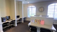 This single-floor space is designed with functionality and accessibility in mind, offering a clean and organized environment suited for community or administrative use. The main area features a prominent reception counter finished with a granite-style surface, serving as a central hub for check-ins and information, as indicated by the sign-in log and displayed materials including a colorful U.S. map. Adjacent to the reception area, a dedicated workstation section lines the wall, equipped with desktop computers and black folding chairs, providing a practical space for computer-based tasks. Decorative columns with classic detailing add architectural character to the room, while crown molding along the ceiling enhances the overall refined aesthetic. The space benefits from generous natural light streaming through an arched window dressed with horizontal blinds, offering a view of the greenery outside. Neutral tones on the walls, paired with dark carpeting, create a balanced and professional atmosphere. A large round wall clock serves as both a functional and decorative accent. The overall ambiance is welcoming, organized, and purpose-driven, making it well-suited for receiving visitors and supporting everyday operational needs.