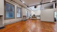 This single-floor unit showcases a compelling mix of historic architectural details and contemporary finishes. Rich, warm hardwood floors run throughout the entire space, lending a sense of continuity and character. The open-plan living and kitchen area is generously proportioned, with high ceilings and exposed industrial-style black piping overhead that reinforce the loft-like aesthetic. Track lighting complements a statement pendant fixture above the kitchen island, providing both functional and ambient illumination.

The living area features two decorative fireplace surrounds flanking a pair of large windows, which allow natural light to fill the space while offering views of the surrounding streetscape. The kitchen is thoughtfully designed with white shaker-style cabinetry, granite countertops, and stainless steel appliances including a range, microwave, refrigerator, and dishwasher. A central island with a built-in sink provides additional workspace and storage. A stacked washer and dryer unit is conveniently integrated into the kitchen area.

A separate room with gray louvered doors offers flexible storage or utility space. The bathroom continues the hardwood flooring and includes a full tub-shower combination with clean, white finishes. The overall ambiance is one of urban character paired with modern functionality.