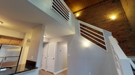This two-floor loft-style space masterfully combines industrial heritage with modern comfort. The main level immediately impresses with dramatic soaring ceilings, exposed brick walls, and rich wood beam ceilings that lend a warm, distinctive character. A white-railed staircase leads to the upper level, with a slatted interior window connecting the two floors and enhancing the open, airy feel.

The kitchen features warm maple cabinetry, dark granite countertops, a peninsula with a breakfast bar, stainless steel refrigerator, and a black range — a functional and stylish space for cooking and entertaining. Light wood-tone flooring flows throughout the main level, complementing the neutral gray walls.

Upstairs, the bedroom offers plush carpeting, dual closets, and the continued warmth of exposed wooden beams and a skylight, filling the room with natural light. A spacious walk-in closet with wire shelving provides generous storage.

The space includes two bathrooms: a full bath with a tub/shower combo and stone-look tile flooring, and a second bath featuring a wood-toned vanity, large mirror, and a well-lit vanity fixture. A convenient louvered-door closet on the main level adds further practicality. Throughout, the interplay of brick, wood, and clean modern finishes creates a compelling and characterful living environment.