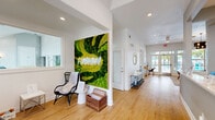 Located at 2603 Silkwood Circle in Orlando, FL, this single-story community clubhouse for Fusion offers a thoughtfully designed and generously sized amenity space. The entry foyer greets visitors with vaulted ceilings, light hardwood flooring, and elegant double glass doors topped by an arched transom window that floods the space with natural light. A staffed reception desk, branded with the Fusion logo and accented by pendant lighting, anchors the front area. Adjacent to reception, a vibrant moss wall art installation bearing the "Fusion" neon sign adds a striking botanical focal point. The expansive living room features stylish pendant chandeliers, a large sectional sofa, and oversized windows and glass doors that open directly to an outdoor pool area, seamlessly blending indoor and outdoor living. A kitchenette with white cabinetry, quartz countertops, a breakfast bar with seating, woven pendant lights, and stainless steel appliances serves the common area. A dedicated children's activity room boasts coffered ceilings, built-in storage in teal, and playful furnishings. The well-equipped fitness center offers high ceilings, rubber flooring, cardio machines, strength equipment, free weights, and colorful acoustic wall panels, providing a complete workout environment.