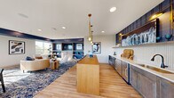Located at 1160 Frost Ave in Maplewood, MN, this thoughtfully designed amenity space delivers a refined, multi-functional environment with a cohesive contemporary aesthetic. The open-concept interior is anchored by warm light wood flooring, bold navy blue accent walls, and recessed lighting complemented by striking brass pendant fixtures that add a touch of elegance throughout.

The lounge area features a built-in shelving and media wall clad in navy patterned tile, providing ample display space alongside a centrally mounted television. Large windows flood the area with natural light and frame outdoor views. Adjacent to the lounge, a dedicated dining area accommodates a sizable wooden table surrounded by upholstered seating, set beneath a sculptural gold pendant light and bordered by expansive windows overlooking the exterior terrace.

The wet bar runs along one wall, outfitted with sleek cabinetry, a herringbone tile backsplash, white countertops, a sink, undercounter refrigeration, and open shelving — ideal for entertaining. A long wooden island provides additional prep and gathering space.

The outdoor terrace extends the living experience with large-format tile pavers, lounge seating, a fire pit, string lights, and a full outdoor grill station, all set against open sky views of the surrounding Maplewood area.