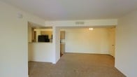 This single-floor space features an open-concept layout that seamlessly connects the living area to the kitchen. The living area is finished with neutral-toned wall-to-wall carpeting, white walls, and a ceiling fan, contributing to a clean and functional atmosphere. A partial dividing wall with a pass-through opening separates the living space from the kitchen, maintaining visual connectivity while defining each area. The kitchen is equipped with light wood cabinetry, a black refrigerator, and additional appliances, all visible through the pass-through. The cabinetry extends along the walls, offering generous storage. HVAC vents are integrated into the ceiling, ensuring climate control throughout the space. The overall design is straightforward and practical, with a neutral color palette of whites and beiges that provides a versatile backdrop. The open flow between the kitchen and living areas makes the space feel cohesive and well-utilized, suitable for everyday living and easy furnishing.