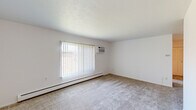 This single-floor unit is designed with a straightforward, functional layout across several distinct spaces. The living area is generously sized, featuring neutral wall-to-wall carpeting, crisp white walls, and a textured ceiling. A large window fitted with vertical blinds allows natural light to fill the room, while a baseboard heating unit and wall-mounted air conditioner ensure year-round climate control.

The bedroom spaces continue the same carpeted flooring and white-painted walls, with one room featuring a ceiling fan with integrated lighting and a window with vertical blinds, complemented by its own baseboard heater. An additional room offers a clean, open feel with ample wall space and neutral carpet.

The galley-style kitchen is efficiently arranged, offering white cabinetry on both sides, light countertops, a stainless steel sink, a dishwasher, a white refrigerator, and a range. Tile flooring runs throughout the kitchen, leading toward a closeted utility area.

The bathroom is compact and well-appointed, featuring tile flooring, a white vanity with under-cabinet storage, a lighted mirror, and a combined tub and shower. The overall ambiance of the unit is clean, neutral, and move-in ready.