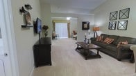 Located at 211 Redwood Dr in Johnstown, Ohio, this single-floor residence offers a thoughtfully arranged living space designed for comfort and functionality. The open-concept layout centers around a generously sized living area finished with plush wall-to-wall carpeting that extends throughout, creating a warm and cohesive feel underfoot. The living room is well-appointed, featuring a wall-mounted flat-screen television, a dark wood media console, and a classic sofa accented with decorative pillows. A wooden coffee table serves as the centerpiece of the seating arrangement, complemented by ambient floor and table lamps that contribute to a welcoming atmosphere. A sliding glass door framed by vertical blinds and a valance-style window treatment allows natural light to filter into the space while providing access to an outdoor area. Beyond the main living area, a secondary sitting or sunroom space is visible, furnished with additional seating and bathed in natural light — ideal for relaxing or reading. The neutral color palette of soft greens and warm whites throughout the walls enhances the sense of openness, while decorative accents and framed artwork add personalized touches. The overall ambiance is cozy, tidy, and move-in ready.