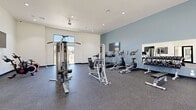 This fitness center offers a generously sized, single-floor exercise room designed to support a comprehensive range of workout routines. The space features soaring vaulted ceilings that lend an open, airy atmosphere, complemented by large windows and glass doors that flood the interior with natural light and provide outdoor views of the surrounding area. A ceiling fan overhead aids in ventilation and comfort during workouts.

The exercise room is thoughtfully organized to accommodate multiple training styles. The cardio section includes a row of treadmills and an elliptical machine, positioned beneath a wall-mounted flat-screen television for entertainment during exercise. Nearby, spin bikes are arranged for cycling workouts. The strength training area is outfitted with multi-function cable machines, a squat/power rack with a bench press, and a dedicated free weights station featuring a full dumbbell rack with mirrors for form monitoring. Additional functional fitness accessories, including stability balls, medicine balls, and resistance bands, are neatly stored along the wall.

The flooring throughout is durable rubber gym tile, practical for both heavy equipment and high-impact activity. The interior walls are finished in a calming slate blue and white color palette, creating a clean and motivating environment. A water station and posted fitness center guidelines near the entrance add practical amenities for users. The space is well-lit with recessed lighting throughout.