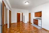 This single-floor unit showcases warm, honey-toned parquet flooring throughout the main living area and bedroom, lending the space a timeless, classic character. The open main room is generously proportioned, bathed in natural light through a large window fitted with an air conditioning unit, and offers views of the surrounding urban streetscape. A set of sliding wood-framed doors opens to a spacious built-in closet complete with shelving and hanging rods, providing ample storage.

The kitchenette is neatly tucked into a dedicated alcove, outfitted with rich wood cabinetry, a white gas range, a sink with dark countertops, and a full-size white refrigerator positioned just outside the alcove for easy access. A separate closet adjacent to the kitchen area adds further storage flexibility.

The bathroom retains a charming vintage aesthetic, featuring classic black-and-white mosaic floor tiles, cream wall tiles accented with bold navy blue tile borders, and a full bathtub with overhead shower. A frosted window above the tub allows for natural ventilation and light. A white vanity completes the space.

The unit's entry is marked by a solid wood door with an arched surround, contributing to the architectural character of the interior. Clean white walls and flush-mount ceiling lighting maintain a bright, functional ambiance throughout.