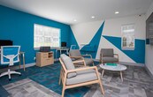 Located at 8855 N River Pkwy in Sandy Springs, Georgia, this single-floor office suite makes a striking first impression with its bold, contemporary design. The space is unified by a vivid teal and white color palette, most dramatically expressed through large geometric graphic murals painted directly onto the walls, creating an energetic and modern atmosphere throughout.

The main area serves a dual purpose, seamlessly blending a functional workstation zone with a comfortable lounge and meeting space. The workstation area features a light wood desk, an ergonomic white mesh chair, and a desktop computer, accompanied by a wooden storage cabinet and printer — providing everything needed for day-to-day office tasks. Natural light streams in through multiple windows fitted with blinds, keeping the space bright and welcoming.

The lounge portion of the suite is furnished with warm-toned wooden armchairs with gray upholstery, a round side table, and a wall-mounted flat-screen television, making it well-suited for informal meetings or relaxed collaboration. A wall-mounted HVAC unit ensures year-round climate comfort.

The flooring throughout is a standout feature — a patchwork carpet tile design blending gray, white, and teal tones that complements the wall graphics and ties the entire space together. A connecting entry area with clean white walls and neutral carpeting provides a calm transition point between rooms, completing this thoughtfully designed professional environment.