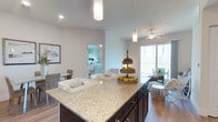 This two-bedroom, two-bathroom residence offers a thoughtfully designed single-floor layout that maximizes both functionality and comfort. The open-concept kitchen serves as the heart of the home, featuring a large granite-topped island with dark cabinetry, providing ample prep and storage space. Elegant pendant lighting hangs above the island, adding a refined touch to the kitchen's contemporary aesthetic.

The kitchen flows seamlessly into the living area, which is bathed in natural light from expansive sliding glass doors that open to an outdoor balcony, offering views of lush greenery. A ceiling fan enhances airflow throughout the shared living space. Rich wood-look flooring runs through the main living areas, adding warmth and cohesion to the design.

A dedicated dining area is visible adjacent to the kitchen, offering a comfortable space for everyday meals or entertaining. The bedrooms are conveniently accessible from the main living space, with one bedroom visible featuring soft, neutral tones and ample room for furnishings. Recessed lighting and neutral wall colors throughout contribute to the bright, clean, and welcoming ambiance of this well-appointed residence.