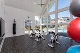 The fitness center at Highlands at Trotters Ridge, located at 2789 Satellite Blvd in Duluth, GA, is a bright and modern single-story amenity space designed to support a well-rounded exercise routine. The room's most striking architectural feature is its dramatic vaulted ceiling with exposed triangular glass panels that rise to a peak, flooding the interior with an abundance of natural light throughout the day. Expansive floor-to-ceiling windows line the primary wall, offering unobstructed views of the resort-style outdoor swimming pool and surrounding lounge area, creating an energizing and visually open atmosphere during workouts. The flooring consists of durable, dark tile laid in a brick pattern, providing both a functional and polished aesthetic. The space is equipped with a row of stationary cycling bikes positioned to face the pool-view windows, making for an immersive and motivating spin experience. Additional fitness equipment includes a heavy punching bag mounted near a full-length wall mirror, a dedicated free-weight and resistance training station stocked with kettlebells, medicine balls, and resistance bands, and open floor space for stretching or functional training. A direct exterior door provides convenient access to the outdoor pool deck, seamlessly connecting indoor fitness activities with the outdoor amenity areas.