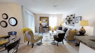 This single-floor space centers around a well-appointed living area that blends comfort and style through a carefully curated mix of furnishings and décor. The sitting area serves as the focal point, anchored by a geometric-patterned area rug in shades of grey, gold, and white that defines the seating arrangement. A tufted accent chair in warm beige, accented with a mustard yellow throw, pairs with a dark rounded lounge chair and a classic striped sofa to create a balanced, inviting conversation area. A circular glass-top coffee table adds an elegant, airy touch at the center.

Natural light fills the room through multiple windows dressed in sheer white and gold curtains, contributing to the bright, open atmosphere. A dedicated workspace is thoughtfully integrated into the background, featuring a wicker chair and a small desk beneath a bold equestrian-themed artwork that adds a striking visual element. Decorative wall-mounted shelving on the opposite wall provides both storage and display opportunities. The textured ceiling and neutral carpet flooring tie the space together, lending warmth and a cozy residential character throughout.