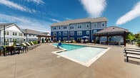 Located at 105 Sweeten Grass Hill in Asheville, NC, this expansive outdoor amenity area at Burton Hills offers a resort-style experience with breathtaking Blue Ridge Mountain views as its backdrop. The centerpiece of the space is a generously sized swimming pool with crystal-clear water, complemented by a separate hot tub for relaxation. The surrounding pool deck is finished in smooth concrete and provides ample room for lounging, with numerous chaise lounge chairs and seating areas thoughtfully arranged throughout. Rustic wooden pergolas add architectural character to the space, offering shaded retreats from the sun. A dedicated outdoor grilling and dining area is also present, making it ideal for social gatherings. Lush landscaping with manicured shrubs and young trees borders the perimeter, adding greenery and a sense of privacy. Adjacent to the pool area is a well-appointed clubhouse building featuring floor-to-ceiling windows and a classic architectural design with a stone chimney, dormer windows, and a standing-seam metal roof. The entire outdoor space is enclosed by a decorative fence and benefits from abundant natural light and open skies, all framed by the stunning panoramic mountain scenery that defines the Asheville region.