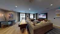 This thoughtfully designed single-floor amenity lounge offers a sophisticated blend of entertainment and relaxation. The main gathering area is anchored by a large wall-mounted flat-screen television paired with a rich wood media console, complemented by an arrangement of plush sectional sofas, upholstered accent chairs, and a rounded ottoman atop a layered area rug — creating an inviting atmosphere for both casual lounging and group viewing. A full-length shuffleboard table adds a recreational focal point, positioned along the perimeter to maximize open floor space. Warm hardwood flooring flows throughout, contrasting beautifully with the cool grey tones of the walls and ceilings. Recessed lighting provides consistent, even illumination, while large windows dressed with floor-length curtains allow generous natural light and offer views to an adjacent outdoor balcony area. Built-in shelving units with curated décor accents add depth and personality to the entertainment zone. A separate dining nook features a sleek glass-top table with contemporary teal accent chairs, ideal for smaller gatherings or meetings. Adjacent to the dining area is a compact kitchenette with dark cabinetry, a countertop sink, and pendant lighting, finished with vibrant artwork. The space is accessible via double entry doors and is appointed with thoughtful details throughout.