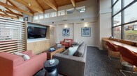 This single-floor space showcases a striking architectural design centered around soaring vaulted ceilings with richly finished exposed wood beams and tongue-and-groove paneling. A continuous band of clerestory windows runs along the upper walls, flooding the interior with natural light and creating an open, airy atmosphere. Large floor-to-ceiling windows along one wall offer views of the surrounding landscape, further enhancing the connection between the indoors and outdoors.

The main lounge area is thoughtfully appointed with a sectional sofa, coral-toned accent chairs, and a wooden coffee table, all arranged around a centrally focused seating layout. A built-in media unit with a wall-mounted flat-screen television and light wood cabinetry anchors one side of the room. A slatted wood partition screen provides subtle division within the open floor plan while maintaining visual continuity throughout the space.

Neutral-toned carpet, warm wall sconces, and carefully selected framed artwork contribute to the refined yet welcoming ambiance. Additional seating areas near the windows offer quieter retreats. The overall design balances natural materials with contemporary furnishings, resulting in a sophisticated and functional communal environment.