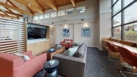 This single-floor space showcases a striking architectural design centered around soaring vaulted ceilings with richly finished exposed wood beams and tongue-and-groove paneling. A continuous band of clerestory windows runs along the upper walls, flooding the interior with natural light and creating an open, airy atmosphere. Large floor-to-ceiling windows along one wall offer views of the surrounding landscape, further enhancing the connection between the indoors and outdoors.

The main lounge area is thoughtfully appointed with a sectional sofa, coral-toned accent chairs, and a wooden coffee table, all arranged around a centrally focused seating layout. A built-in media unit with a wall-mounted flat-screen television and light wood cabinetry anchors one side of the room. A slatted wood partition screen provides subtle division within the open floor plan while maintaining visual continuity throughout the space.

Neutral-toned carpet, warm wall sconces, and carefully selected framed artwork contribute to the refined yet welcoming ambiance. Additional seating areas near the windows offer quieter retreats. The overall design balances natural materials with contemporary furnishings, resulting in a sophisticated and functional communal environment.