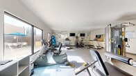 This fitness center offers a comprehensive, single-level workout facility thoughtfully designed to accommodate a wide range of exercise needs. The main cardio and strength area is bright and open, benefiting from large windows that invite natural light and provide outdoor views, creating an energizing atmosphere. The space is equipped with multiple treadmills, ellipticals, stair climbers, stationary bikes, and a full rack of dumbbells, complemented by strength training machines. A bold, typographic feature wall adorned with fitness-themed words in teal and grey serves as both a visual focal point and motivational backdrop, with wall-mounted flat-screen TVs positioned for convenient viewing during workouts. Large-format tile flooring throughout lends a clean, modern aesthetic. An adjoining studio room provides a dedicated space for stretching, yoga, or group exercise, featuring full-length mirrors, a wall-mounted ballet barre, wood-look tile flooring, its own flat-screen TV, and climate control. Open shelving units offer convenient storage for personal items. Recessed lighting throughout ensures consistent illumination across all areas. The overall design is contemporary and functional, delivering a well-rounded fitness environment suited to users of all levels.