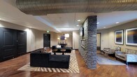 This sophisticated single-floor space makes a strong first impression with its thoughtfully designed interior and blend of modern and industrial elements. The ceiling draws the eye upward, where exposed corrugated metal ductwork adds an industrial character alongside recessed lighting and drop-ceiling panels, creating a layered and dynamic overhead aesthetic. Hardwood flooring in a rich warm tone runs throughout, anchoring the space with warmth and elegance. A standout architectural feature is the floor-to-ceiling stacked stone column that houses a sleek linear fireplace, serving as a dramatic focal point and natural divider within the open layout. The lounge area is furnished with plush, low-profile dark sofas arranged atop a bold zebra-print area rug, complemented by a round wooden side table and an arc floor lamp that adds ambient lighting. A console table styled with framed artwork and decorative accents occupies the background, contributing to the curated, upscale atmosphere. Additional seating areas are visible beyond the stone column, suggesting a spacious and versatile layout. Neutral beige walls and subtle recessed lighting throughout maintain a refined, welcoming ambiance ideal for relaxation or social gathering.