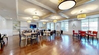 Located at 1715 Tamiami Trail in Bradenton, Florida, this single-floor dining and kitchen space offers a welcoming and well-appointed environment designed for communal dining and gathering. The main dining area is distinguished by its elegant coffered ceilings adorned with decorative molding and a series of stylish drum pendant light fixtures that provide warm, even illumination throughout the room. Rich, warm-toned hardwood flooring runs seamlessly across the entire space, adding a classic and inviting character. Multiple dining tables with mixed seating — including cushioned chairs — are thoughtfully arranged to accommodate a variety of group sizes. A built-in shelving and cabinet unit along the back wall, paired with a mounted flat-screen television, adds both functionality and a social focal point to the dining area. A separate seating nook near the entrance benefits from generous natural light through large windows and glass double doors, creating a bright and airy atmosphere with views to the outdoors. The fully equipped kitchen features rich dark wood cabinetry, granite countertops, a mosaic tile backsplash, a central peninsula, and stainless steel appliances including a refrigerator, wall oven, and microwave. Pendant lighting above the peninsula and recessed ceiling lights complete the kitchen's polished, functional design.