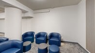 This well-appointed single-floor space offers a thoughtfully designed open layout that accommodates multiple functional zones within a generous footprint. The interior features a cohesive modern aesthetic defined by crisp white walls accented by a bold navy blue feature wall, patterned carpet tile flooring, and light wood-tone hardwood in the kitchenette area.

A central communal high-top table with matching navy blue chairs serves as the primary gathering and collaboration area, ideal for group work or casual meetings. Adjacent to this, a dedicated lounge nook provides a more relaxed setting, furnished with rounded blue leather armchairs and low cylindrical ottomans and side tables, creating an inviting social corner. A mounted flat-screen display and slatted wood accent wall define a media or presentation alcove near the lounge area.

The kitchenette is equipped with a stainless steel refrigerator, upper cabinetry, and counter space, supporting day-to-day convenience. Exposed conduit along the ceiling adds an industrial touch, while flush-mounted ceiling lights ensure consistent, even illumination throughout. A wall-mounted mini-split unit provides climate control. A vibrant abstract artwork displayed on the main wall adds a dynamic focal point, contributing to the space's energetic yet functional character.