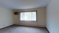 This single-floor space offers a clean, straightforward layout designed for comfortable everyday living. The main living area is finished with wall-to-wall carpeting that adds warmth underfoot and contributes to a quiet, cozy atmosphere. Neutral-toned walls keep the space feeling bright and open, providing a versatile backdrop for a variety of furnishing styles.

A prominent large window fitted with vertical blinds serves as the focal point of the main room, allowing generous natural light to filter in while offering views of mature trees outside — a pleasant natural backdrop that brings a sense of the outdoors inside. The vertical blinds provide flexible light and privacy control throughout the day.

Climate comfort is well-addressed, with a wall-mounted air conditioning unit installed for warm-weather cooling and a baseboard heating system running along the base of the exterior wall to provide efficient warmth during colder months. This dual climate-control setup ensures year-round comfort.

The overall ambiance of the space is understated and functional, with a neutral palette and practical fixtures that make it easy to personalize. The single-floor layout offers convenient, step-free accessibility throughout, making it a practical and adaptable space suitable for a range of occupants.