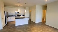 This single-floor residence offers a clean, contemporary design with an open-concept layout that seamlessly connects the kitchen and living areas. The kitchen is a standout feature, showcasing crisp white cabinetry that extends to the upper walls, bold dark countertops, and a curved island with a built-in sink — ideal for both meal preparation and casual dining. Stainless steel appliances, including a range, microwave, and additional built-in units, lend a sleek, modern finish. Pendant lighting hangs above the island, complemented by a track lighting fixture that illuminates the workspace with a warm, functional glow. The flooring throughout is a rich hardwood-style material in a warm grey-brown tone, adding warmth and continuity across the open space. The neutral white walls and light-filled interior create a bright, airy ambiance. A hallway visible from the main area leads to additional rooms, including a bathroom, suggesting a well-organized and practical floor plan. The overall aesthetic is modern and move-in ready, combining stylish finishes with functional design elements throughout.