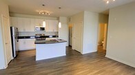 This single-floor residence offers a clean, contemporary design with an open-concept layout that seamlessly connects the kitchen and living areas. The kitchen is a standout feature, showcasing crisp white cabinetry that extends to the upper walls, bold dark countertops, and a curved island with a built-in sink — ideal for both meal preparation and casual dining. Stainless steel appliances, including a range, microwave, and additional built-in units, lend a sleek, modern finish. Pendant lighting hangs above the island, complemented by a track lighting fixture that illuminates the workspace with a warm, functional glow. The flooring throughout is a rich hardwood-style material in a warm grey-brown tone, adding warmth and continuity across the open space. The neutral white walls and light-filled interior create a bright, airy ambiance. A hallway visible from the main area leads to additional rooms, including a bathroom, suggesting a well-organized and practical floor plan. The overall aesthetic is modern and move-in ready, combining stylish finishes with functional design elements throughout.