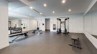 Located at 1650 Ferry Wharf Rd in Kelowna, BC, this fitness facility offers a comprehensively equipped exercise room and connecting hallway across a single floor. The space is designed with a clean, modern aesthetic, featuring neutral white walls, recessed ceiling lighting, and durable dark flooring that contributes to a professional gym atmosphere.

The exercise room is generously sized, accommodating a wide variety of fitness equipment across distinct zones. The cardio area is lined with multiple treadmills, an elliptical trainer, a stair climber, and a mini trampoline, providing ample options for cardiovascular workouts. Large mirrors along the walls visually expand the space and allow users to monitor their form. A dedicated free weights section features a well-organized dumbbell rack with a full range of weights, accompanied by adjustable benches. The strength training area includes a cable machine, additional benches, and a wall-mounted storage rack for accessories.

Natural light filters in through windows on one side of the room, offering pleasant outdoor views of the surrounding greenery. A wall-mounted air conditioning unit ensures climate comfort year-round. A built-in water fountain provides added convenience. One wall is accented with warm wood slat paneling, adding a refined design detail that contrasts tastefully with the otherwise crisp, minimalist interior. The hallway provides straightforward access into the main exercise area.