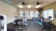 This fitness center offers a comprehensive workout environment designed to support a wide range of exercise routines. The space features durable dark rubber flooring throughout, providing a safe and stable surface for all types of physical activity. Warm neutral-toned walls and flush-mounted fluorescent lighting create a bright, energizing atmosphere, while two ceiling fans ensure consistent air circulation during workouts.

Natural light filters in through multiple windows fitted with horizontal blinds, offering views of the surrounding outdoors and adding a refreshing openness to the space. The cardio section is well-appointed with Life Fitness elliptical trainers, a treadmill, and a recumbent bike, catering to a variety of cardiovascular fitness preferences. A wall-mounted flat-screen television provides entertainment options for users during their sessions.

The strength training area features a multi-function cable machine and a weight bench, enabling users to perform a broad spectrum of resistance exercises. Yoga or exercise mats are also available on the floor for stretching and floor-based workouts. A water fountain is conveniently accessible along the wall, and ample open floor space throughout ensures comfortable movement between equipment. This fitness center is thoughtfully laid out to accommodate multiple users simultaneously.