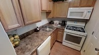 This single-story space includes a well-organized kitchen that serves as a central feature. The kitchen is outfitted with light natural wood cabinetry that lines both the upper and lower walls, providing ample storage throughout. The countertops display a granite-style laminate surface with warm brown and tan tones, offering generous prep space along an L-shaped layout. White appliances include a gas range with oven, an over-the-range microwave, and a dishwasher, creating a cohesive and clean aesthetic. A stainless steel sink is set into the countertop at the corner junction of the L-shape, positioned beneath upper cabinetry for added convenience. The flooring throughout is warm-toned hardwood, which complements the light wood cabinetry and contributes to an inviting, cohesive atmosphere. The overall design balances practicality with a classic, approachable aesthetic, making the kitchen a comfortable and efficient workspace suited for everyday use.
