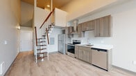 Located in Bellingham, WA, this two-story loft-style unit at 1207 Harris Ave, Unit 306 offers a thoughtfully designed layout spread across two floors. The main level features an open-concept kitchen and living area with soaring ceilings that create a striking sense of vertical space. The kitchen is well-equipped with stainless steel appliances, including an electric range with a stainless steel hood vent, a refrigerator, and a dishwasher. Taupe-toned shaker-style cabinetry with brushed nickel hardware pairs with white countertops, offering both style and functionality. Light wood-tone flooring runs throughout, lending warmth and continuity to the space. A sleek open-riser staircase with a rich wood-and-white metal design serves as both a functional and architectural focal point, connecting the two floors. Large windows on the upper level frame views of the surrounding Bellingham neighborhood, filling the space with natural light. The upper floor provides additional living space with the same light flooring. The bathroom features a full tub-shower combination, a single vanity with white countertops and wood cabinetry, and clean, bright finishes throughout. An accessible electrical panel is conveniently integrated into the layout. This unit combines modern finishes with a dynamic two-level design, ideal for those seeking a contemporary urban living experience.