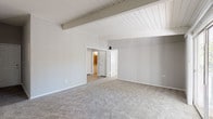 Located at 3214 Valley Bluff Dr in Atlanta, GA 30340, this single-story home showcases a consistent architectural character defined by its striking white-painted wood beam and plank ceilings, which lend a mid-century warmth throughout the living spaces. Neutral gray walls and plush wall-to-wall carpeting flow through the spacious living room and bedrooms, creating a cohesive and understated palette. The living room is a standout space, featuring a sliding glass door that opens directly onto a covered deck with views of surrounding greenery, allowing natural light to fill the room. The kitchen is equipped with white cabinetry, ample upper and lower storage, a double sink, and black appliances including a range and dishwasher, all set over durable wood-look flooring. The bedrooms are generously sized, each benefiting from the signature beamed ceilings, carpeted floors, and good natural light. One bedroom features open shelving along an entire wall, offering convenient storage. The bathroom is well-appointed with a wide vanity featuring a stone-look countertop, white cabinetry, a large mirror, and a tub/shower combination fully tiled in warm beige. The home's layout is practical and open, with a comfortable flow between rooms suitable for everyday living.