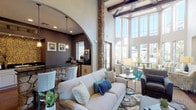 This well-appointed communal space showcases an impressive architectural design that blends rustic and contemporary elements throughout. The centerpiece of the main lounge area is a dramatic soaring ceiling accented by exposed wooden beams and striking natural stone columns that anchor the room with a warm, lodge-inspired character. Floor-to-ceiling windows along the exterior wall allow an abundance of natural light to pour in while framing views of the surrounding outdoor landscape, including manicured greenery and palm trees.

The lounge is furnished with a thoughtfully curated mix of comfortable seating, including plush sofas, upholstered armchairs, and accent chairs arranged around area rugs in teal and earth tones. A media wall features floating shelves and a mounted flat-screen television, creating a cozy gathering spot. Decorative elements such as ceramic lamps, vases, and artwork contribute to the cohesive teal, blue, and neutral color palette.

An elegant arched stone entryway leads to a dedicated kitchenette area, finished with dark granite countertops, white cabinetry, polished concrete floors, and stainless steel appliances including a coffee station. The kitchenette is well-equipped for hosting and entertaining. Throughout the space, rich hardwood flooring ties the areas together, complementing the natural stone and beam details for a refined yet welcoming ambiance.
