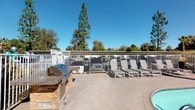 This single-story residential community features a well-appointed outdoor amenity area and a centrally located mailroom hub designed for everyday convenience. The outdoor space includes a spacious concrete patio with a built-in BBQ grill station set atop a stacked stone base with a granite countertop, ideal for outdoor entertaining. A row of sling-style lounge chairs lines the patio, offering a relaxing area framed by mature trees and distant mountain views beneath open, sunny skies. The space is enclosed by a white metal fence that provides both security and an unobstructed view of the surrounding landscape. The covered mailroom area is conveniently accessible from the main walkway and features two full banks of individual U.S. Mail letter boxes mounted along the wall, accommodating a large number of residents. A dedicated parcel drop box is also present. The structure features light-toned horizontal lap siding with a neutral palette that blends seamlessly into the broader community aesthetic. Wide, sun-drenched walkways connect the mailroom to the rest of the property, with manicured landscaping visible throughout. The overall ambiance is practical, clean, and community-oriented, offering residents easy access to essential services in a well-maintained outdoor setting.