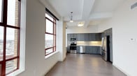 This single-floor space combines an open living area with a well-appointed kitchen, creating a cohesive and functional layout. The kitchen is a standout feature, outfitted with slate-gray shaker-style cabinetry that extends along two walls, offering substantial storage. Stainless steel appliances, including a range, microwave, dishwasher, and refrigerator, lend a polished, contemporary feel. A decorative tile backsplash adds visual texture behind the range, complementing the light-toned countertops. Pendant and flush-mount ceiling fixtures provide focused task lighting throughout the kitchen area. The open living space transitions seamlessly from the kitchen, finished with wide-plank light wood flooring that runs throughout, contributing to a clean and airy aesthetic. A large, multi-pane window with warm wood-toned frames floods the space with natural light and frames an urban cityscape, offering an elevated view of the surrounding neighborhood. The tray ceiling detail above the kitchen adds architectural dimension, while the overall neutral color palette of whites and grays creates a calm, modern atmosphere well-suited for both everyday living and entertaining.