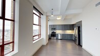 This single-floor space combines an open living area with a well-appointed kitchen, creating a cohesive and functional layout. The kitchen is a standout feature, outfitted with slate-gray shaker-style cabinetry that extends along two walls, offering substantial storage. Stainless steel appliances, including a range, microwave, dishwasher, and refrigerator, lend a polished, contemporary feel. A decorative tile backsplash adds visual texture behind the range, complementing the light-toned countertops. Pendant and flush-mount ceiling fixtures provide focused task lighting throughout the kitchen area. The open living space transitions seamlessly from the kitchen, finished with wide-plank light wood flooring that runs throughout, contributing to a clean and airy aesthetic. A large, multi-pane window with warm wood-toned frames floods the space with natural light and frames an urban cityscape, offering an elevated view of the surrounding neighborhood. The tray ceiling detail above the kitchen adds architectural dimension, while the overall neutral color palette of whites and grays creates a calm, modern atmosphere well-suited for both everyday living and entertaining.