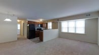 This single-floor space showcases an open-concept layout that seamlessly connects the kitchen and living areas, creating a cohesive and functional living environment. The kitchen is outfitted with rich, warm-toned wood cabinetry that provides ample storage, complemented by dark countertops and a full suite of black appliances, including a refrigerator, range, and over-the-range microwave. A raised peninsula serves as a natural divider between the kitchen and the living space, offering a convenient spot for casual dining or meal prep while maintaining the open feel of the floor plan. The kitchen also features hardwood-style flooring, adding a practical and visually appealing contrast to the rest of the space. The living area is finished with plush, neutral-toned carpeting that adds warmth and comfort underfoot. A large, multi-pane window floods the living space with natural light, enhancing the bright and airy ambiance of the room. The neutral wall tones throughout create a clean, versatile backdrop suitable for a variety of interior styles. A hallway visible toward the rear of the unit suggests additional private rooms beyond the main living area. Overall, the space offers a practical and comfortable layout with a welcoming atmosphere.