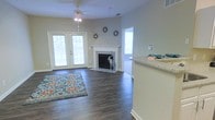 Located at 1000 Henrico Ln in Morrisville, NC, this single-floor residence offers a well-designed, open layout that balances comfort and functionality. The living area serves as the heart of the home, highlighted by rich, dark hardwood-style flooring that runs throughout the space, adding warmth and a polished aesthetic. A classic white-mantled fireplace serves as a natural focal point, flanked by decorative accents that contribute to the room's inviting character. A ceiling fan overhead ensures year-round comfort. Natural light floods the interior through a set of French doors, which provide a seamless connection to the outdoors while offering views of the surrounding exterior. An additional window further brightens the space, enhancing the open and airy feel. Adjacent to the living area, a partial view of the kitchen reveals granite-style countertops, hinting at quality finishes carried throughout the home. The neutral wall tones provide a versatile backdrop that complements a variety of décor styles. Overall, this residence presents a cohesive and comfortable living environment with thoughtful design details, abundant natural light, and a practical single-level floor plan well-suited for everyday living.