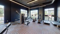 This fitness center offers a thoughtfully designed workout environment spread across a single level. The space is defined by its bold navy blue walls, exposed industrial ceiling elements, and linear LED lighting that runs along the upper perimeter, creating a sleek, modern atmosphere. Patterned carpet tile flooring throughout adds a refined yet durable finish suited to high-traffic use.

Large floor-to-ceiling windows line multiple walls, flooding the interior with abundant natural light and providing open views of the surrounding outdoor greenery and streetscape. The generous ceiling height enhances the sense of spaciousness throughout.

Equipment is organized across distinct zones. The cardio area features treadmills, ellipticals, a stair climber, and stationary bikes, including a Peloton cycle, all positioned to take advantage of the natural light and exterior views. The strength training zone includes a cable machine and a full power rack with a bench press station. A dedicated free weights area offers a complete dumbbell rack and a set of kettlebells on a compact stand. Open shelving along one wall provides accessible storage for fitness accessories. Full-length mirrors in the strength zone allow users to monitor their form. The space is accessible via a glass-paneled entry door.