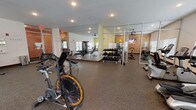 The fitness center at Victory Knoll is a thoughtfully designed, single-floor facility that provides a full range of workout options for residents. The main exercise area features durable rubber flooring throughout and is lined with large wall-to-wall mirrors that enhance the sense of openness. Recessed lighting and ceiling fans ensure a comfortable environment during workouts.

The cardio section is generously stocked with treadmills, ellipticals, upright and recumbent bikes, and rowing machines, all positioned in front of windows with natural light filtering through slatted blinds. Multiple wall-mounted flat-screen televisions are positioned throughout, offering entertainment during exercise. A dedicated strength training zone includes a multi-function cable machine, adjustable weight benches, and a full rack of dumbbells for free weight training.

A separate spin studio provides an intimate space for cycling workouts, distinguished by its warm wood-paneled accent wall and a mounted flat-screen TV. This room is outfitted with several stationary spin bikes and a well-stocked rack of fitness accessories including exercise balls, resistance bands, and yoga mats. A ceiling fan completes the comfortable atmosphere of this dedicated space. Together, the areas create a versatile and well-appointed fitness destination.