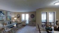 This single-floor residence offers a thoughtfully arranged living space with a warm, neutral color palette that creates a calm and inviting atmosphere throughout. The open-concept layout connects the living and dining areas seamlessly, promoting a natural flow between spaces.

The living area is carpeted and bathed in natural light, courtesy of multiple windows dressed with white horizontal blinds. Decorative wall art, including large starburst-style medallions, adds a stylish touch to the walls, complementing the understated beige tones. A glass-top coffee table and accent chairs with patterned upholstery contribute to the space's refined yet approachable aesthetic.

A console table with slender metallic legs is positioned beneath one of the windows, styled with decorative vases and glass accents that catch the light. The textured ceiling adds subtle architectural character to the room.

Adjacent to the living area, a glimpse into the dining space reveals additional seating and continued use of natural light, with what appears to be access to an outdoor area or enclosed porch. This connection to the exterior enhances the overall sense of openness. The space is well-suited for comfortable everyday living, with its cohesive design choices and functional layout making it both practical and visually appealing.