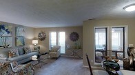 This single-floor residence offers a thoughtfully arranged living space with a warm, neutral color palette that creates a calm and inviting atmosphere throughout. The open-concept layout connects the living and dining areas seamlessly, promoting a natural flow between spaces.

The living area is carpeted and bathed in natural light, courtesy of multiple windows dressed with white horizontal blinds. Decorative wall art, including large starburst-style medallions, adds a stylish touch to the walls, complementing the understated beige tones. A glass-top coffee table and accent chairs with patterned upholstery contribute to the space's refined yet approachable aesthetic.

A console table with slender metallic legs is positioned beneath one of the windows, styled with decorative vases and glass accents that catch the light. The textured ceiling adds subtle architectural character to the room.

Adjacent to the living area, a glimpse into the dining space reveals additional seating and continued use of natural light, with what appears to be access to an outdoor area or enclosed porch. This connection to the exterior enhances the overall sense of openness. The space is well-suited for comfortable everyday living, with its cohesive design choices and functional layout making it both practical and visually appealing.