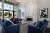 Located at 11972 W Fairview Ave in Boise, Idaho, this single-floor space serves as a polished leasing and community lounge facility. The interior is immediately distinguished by its soaring double-height ceilings, which lend an expansive, airy atmosphere throughout the main lounge area. Floor-to-ceiling windows with black-framed grid detailing line the exterior walls, flooding the space with natural light and offering clear views of the surrounding outdoor environment.

The central lounge is anchored by a striking floor-to-ceiling marble accent wall featuring dramatic veining in cream and gold tones, housing a sleek linear fireplace at its base. A modern circular chandelier hangs overhead, complementing the refined aesthetic. Seating throughout the lounge consists of coordinated navy blue sofas and accent chairs arranged around a gold-framed glass coffee table atop a geometric patterned area rug, all set over warm-toned hardwood flooring.

Adjacent to the lounge, a welcoming reception area includes a dark wood credenza, a wall-mounted television, and a coffee station. Private consultation rooms, visible through interior windows, are furnished for one-on-one meetings. The overall ambiance is contemporary and welcoming, balancing professional functionality with stylish, comfortable design elements.