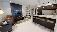 This single-floor social lounge is designed with a contemporary aesthetic, blending functional amenities with stylish interior elements. The space is divided into two distinct zones that flow seamlessly into one another. The lounge area features a large wall-mounted flat-screen television set against a bold dark accent wall, creating a striking focal point. Comfortable seating, including low-profile sofas and ottomans arranged over a textured area rug, provides an inviting setting for relaxed conversation or entertainment. A geometric, diamond-pattern open shelving unit adds a decorative architectural element, displaying curated objects and complementing the modern design language throughout.

Adjacent to the lounge, a kitchen-style bar counter extends along one side of the space, topped with a clean white surface and lined with industrial-style wood and metal bar stools. Dark cabinetry overhead and a built-in oven contribute to a fully appointed feel. Pendant lighting above the counter adds warmth. The bright white tile flooring throughout enhances the sense of openness, while recessed ceiling lighting ensures the space remains well-lit and welcoming.