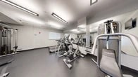 Located at 3232 Eastern Ave in Baltimore, MD, this fitness center offers a compact yet thoughtfully designed workout space across a single floor. The room features a clean, modern aesthetic with white walls, a smooth drop ceiling with recessed rectangular fluorescent lighting, and durable rubber-textured flooring in a dark speckled finish — practical and well-suited for an active environment. Large floor-to-ceiling mirrors line one wall, amplifying the sense of space and allowing users to monitor their form during exercise. The cardio section is outfitted with multiple treadmills and elliptical trainers, as well as a stationary bike, all arranged to maximize the available floor area. A multi-station strength machine and free weight bench provide options for resistance training, complemented by a small collection of dumbbells. A wall-mounted flat-screen television adds convenience for entertainment during workouts. A built-in water station with dual drinking fountains and a bottle-filling feature serves practical hydration needs. Natural light filters in through a window on one wall, and a dedicated entry door connects the fitness center to the broader building. The overall ambiance is functional and well-maintained, delivering an efficient and comfortable facility for daily exercise.