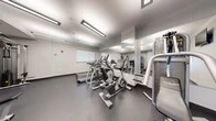 Located at 3232 Eastern Ave in Baltimore, MD, this fitness center offers a compact yet thoughtfully designed workout space across a single floor. The room features a clean, modern aesthetic with white walls, a smooth drop ceiling with recessed rectangular fluorescent lighting, and durable rubber-textured flooring in a dark speckled finish — practical and well-suited for an active environment. Large floor-to-ceiling mirrors line one wall, amplifying the sense of space and allowing users to monitor their form during exercise. The cardio section is outfitted with multiple treadmills and elliptical trainers, as well as a stationary bike, all arranged to maximize the available floor area. A multi-station strength machine and free weight bench provide options for resistance training, complemented by a small collection of dumbbells. A wall-mounted flat-screen television adds convenience for entertainment during workouts. A built-in water station with dual drinking fountains and a bottle-filling feature serves practical hydration needs. Natural light filters in through a window on one wall, and a dedicated entry door connects the fitness center to the broader building. The overall ambiance is functional and well-maintained, delivering an efficient and comfortable facility for daily exercise.