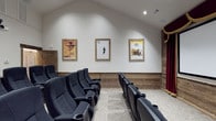 This single-floor home theater is thoughtfully designed to deliver an authentic cinematic experience in a private setting. The screening room features rows of plush, black cinema-style seats complete with built-in cup holders, arranged to provide clear sightlines to the screen. The seating is divided into two sections by a central aisle, accommodating a generous number of guests for a truly communal viewing experience.

The room's architectural design includes vaulted ceilings that lend an open, airy quality to the space, while recessed lighting along the ceiling provides soft, ambient illumination. The lower portion of the walls is clad in warm wood paneling, adding a rustic yet refined character to the room, while the upper walls are finished in clean white, keeping the focus on the screen and décor.

Adding to the cinematic charm, three framed classic Western movie posters—each set in distressed wood frames—are displayed prominently along the side wall, evoking a nostalgic nod to Hollywood's golden era. Rich red curtain draping is visible along one side, further reinforcing the classic movie-house aesthetic. The overall ambiance strikes a balance between the warmth of a home environment and the immersive atmosphere of a professional cinema.