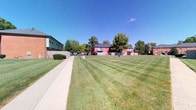 This single-story property is distinguished primarily by its generous outdoor grounds, which serve as a central feature of the space. The exterior showcases a broad, meticulously maintained lawn with freshly mowed striped patterns, reflecting attentive upkeep and offering a welcoming open-air environment. A smooth concrete walkway runs along the perimeter, providing clear and convenient pedestrian access throughout the grounds.

Mature deciduous trees are positioned around the property, offering natural shade and contributing to a pleasant, established landscape. A wood privacy fence defines the boundaries of the outdoor area, creating a sense of enclosure while maintaining a neighborly aesthetic. The surrounding environment includes adjacent brick residential buildings and additional greenery, reinforcing the communal character of the setting.

The outdoor space benefits from abundant natural light under open skies, lending an airy and well-lit ambiance throughout the day. Overall, the property's exterior presents a clean, organized, and functional outdoor environment suited for everyday use and community activity.