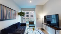 Located at 1201 N Sandburg Terrace in Chicago's prestigious Gold Coast neighborhood, this single-floor residence offers a well-appointed layout with modern finishes throughout. The living area is anchored by a tufted navy blue sofa and a patterned area rug, centered around a large sliding window that frames views of the surrounding urban streetscape, flooding the room with natural light. A sleek media console with a wall-mounted television completes the space.

The kitchen features light wood lower cabinetry contrasted by slate-blue upper cabinets and open white shelving. A textured tile backsplash, matte black fixtures, and under-cabinet lighting add refined detail, while the light hardwood flooring extends seamlessly throughout the unit.

The bedroom is a calm, well-lit retreat with large windows offering street-level city views, light hardwood floors, and soft neutral tones. A connected closet provides practical storage.

The bathroom is finished with a dark wood vanity, white quartz countertop, a large mirror, and a tub-shower combination accented by horizontal tile banding in warm earth tones, giving the space a clean, contemporary feel.