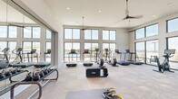This single-floor amenity space is divided into two distinct areas: a well-appointed fitness center and a sky lounge, both characterized by a clean, contemporary design aesthetic. The fitness center commands attention with its soaring double-height ceilings and expansive black-framed windows that span nearly the full height of the walls, flooding the space with natural light and offering views of an outdoor pool area and surrounding landscape. Ceiling fans suspended from the elevated ceiling enhance airflow throughout the workout area. The fitness floor features herringbone-patterned carpet and is equipped with a comprehensive selection of cardio machines, including treadmills, ellipticals, and stationary bikes, alongside strength training equipment such as kettlebells, functional trainers, and sled-style fitness sleds. Full-length mirrors along one wall visually expand the space further. A large-format television is mounted within a geometric tile accent wall, accompanied by built-in storage cubbies. Adjacent to the fitness center, the sky lounge offers a welcoming retreat with warm hardwood-style flooring, white walls adorned with contemporary artwork, and comfortable seating arrangements. Large windows continue the theme of abundant natural light, creating a bright, open atmosphere throughout.