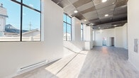 This single-floor space offers a thoughtfully designed layout that blends modern finishes with an open, airy atmosphere. The main open area is defined by soaring ceiling heights and a row of large, floor-to-ceiling black-framed windows that flood the interior with abundant natural light, casting dramatic sun patterns across the light-toned laminate flooring. The exposed industrial-style ceiling, featuring metallic grid panels with recessed lighting, adds a loft-like character throughout the primary areas.

The fully equipped kitchen is a standout feature, showcasing gray shaker-style cabinetry with ample upper and lower storage, white countertops, a stainless steel sink, and white appliances including a refrigerator and range. Exposed brick-painted walls lend additional texture and character to the kitchen area, which also benefits from its own elevated ceiling height.

Multiple private rooms provide flexible use options, each featuring the same light laminate flooring, clean white walls, and windows that bring in natural light and neighborhood views. Baseboard heating is present throughout, ensuring year-round comfort.

The bathroom is finished with large-format, marble-look porcelain tile in the walk-in shower, complemented by chrome grab bars and a clean white vanity. The overall palette of whites and light neutrals creates a cohesive, contemporary feel throughout the entire space.